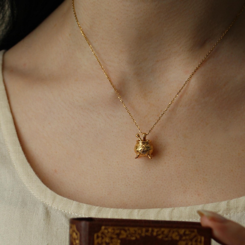 Puede incluir: Collar dorado con un colgante de caldero. El colgante tiene detalles intrincados y peque&ntilde;as patas. El collar se lleva puesto, con la cadena visible. El fondo es de un color suave y neutro. El collar es de oro.