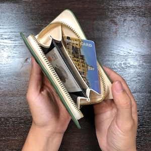 May include: A green and cream-coloured zipped wallet, open to show a ten-pound note and a library card. The wallet has a gold zip and is held open, showcasing its interior compartments. The wallet is made of a smooth material.