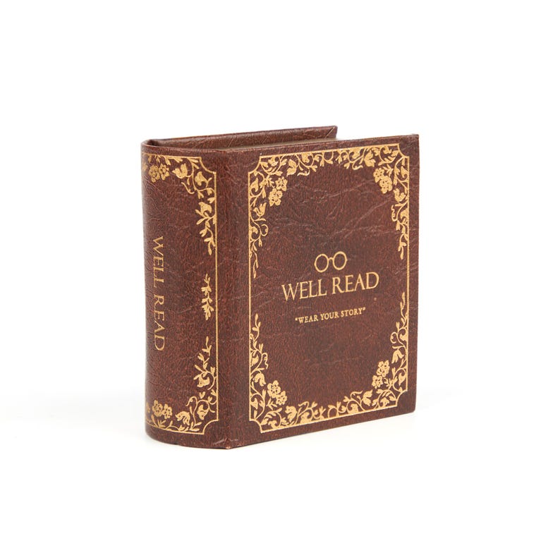 Puede incluir: Una cartera marr&oacute;n con forma de libro con detalles florales dorados y las palabras "WELL READ" y "WEAR YOUR STORY". La cartera est&aacute; de pie y parece estar hecha de cuero o un material similar.