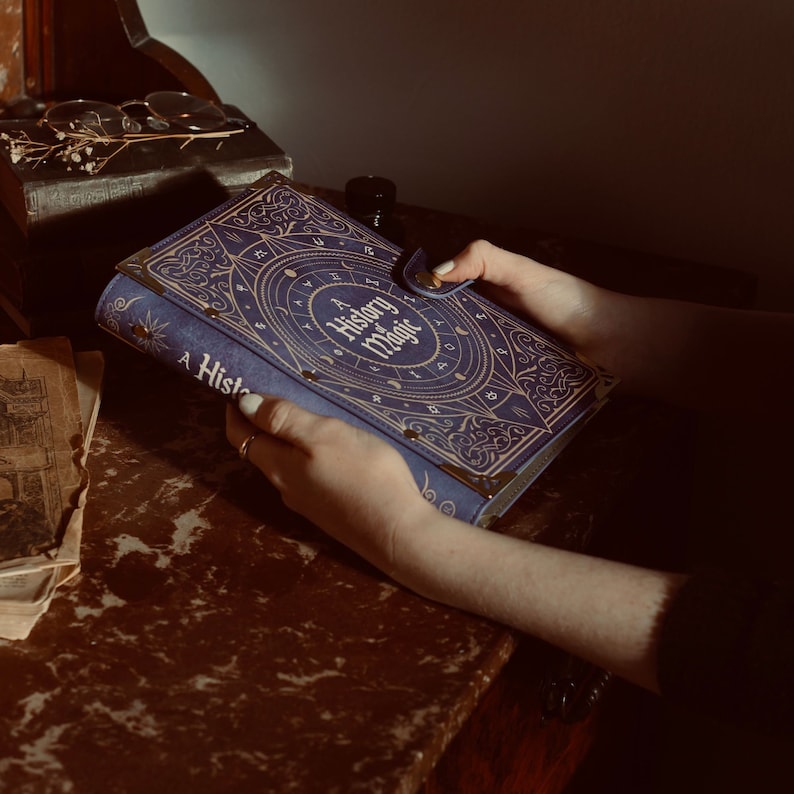May include: A blue leather-bound journal with gold accents and the title "A History of Magic" on the cover. The journal is being held by a person's hands.