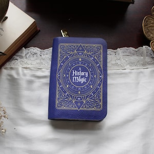 May include: A deep blue wallet in the shape of a book, embossed with gold-coloured designs and the text "A History of Magic". The wallet has a gold-coloured zip and is lying on a white surface. The design is reminiscent of an antique book.