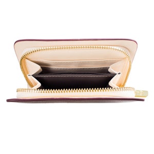 May include: Cream-coloured wallet with a burgundy trim and a gold zip, open to show the interior. The inside has multiple compartments and card slots, with a dark brown lining. Designed for carrying cards and cash.