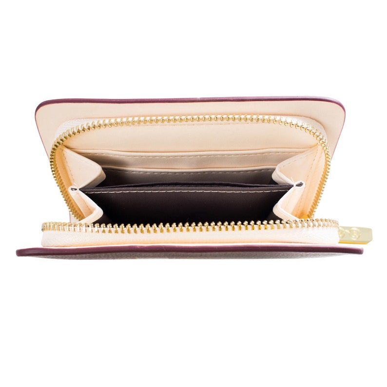 May include: Cream-coloured wallet with a burgundy trim and a gold zip, open to show the interior. The inside has multiple compartments and card slots, with a dark brown lining. Designed for carrying cards and cash.