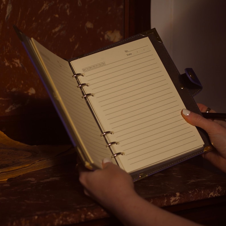 May include: A brown leather bound notebook with gold rings and a cream colored lined paper interior. The notebook is open and the pages are blank. The notebook has the words "No." and "Date" printed on the top right corner.