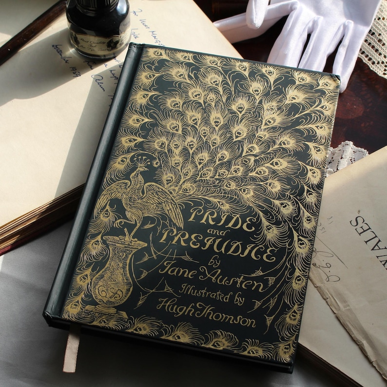 May include: A hardcover book titled "Pride and Prejudice" by Jane Austen, illustrated by Hugh Thomson. The cover is dark teal with gold peacock feather and bird illustrations. A white ribbon bookmark is visible.