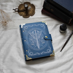 May include: A blue journal with a decorative sword and dragon design on the cover. The journal has gold corner accents and a snap closure. Other items in the image include a brass candle holder, inkwell, and books.