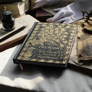May include: A hardcover book titled "Pride and Prejudice" by Jane Austen, illustrated by Hugh Thomson. The cover is black with a gold peacock feather design. An inkwell, pen, and other vintage items are in the background.