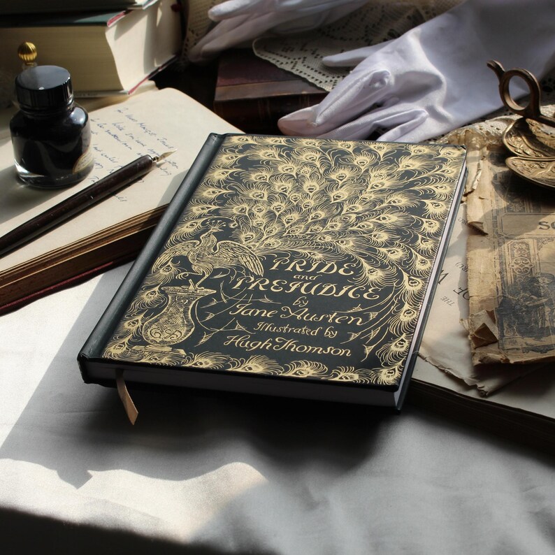 May include: A hardcover book titled "Pride and Prejudice" by Jane Austen, illustrated by Hugh Thomson. The cover is black with a gold peacock feather design. An inkwell, pen, and other vintage items are in the background.