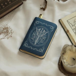 Op de afbeelding: Een blauwe, boekvormige portemonnee met een gouden rits en een decoratief zwaard- en schildontwerp. De portemonnee ligt op een wit oppervlak, met een kaars en een open boek op de achtergrond.