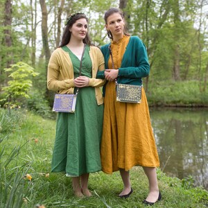 May include: Two women wearing dresses and cardigans stand in a woodland setting. One woman is wearing a green dress and a yellow cardigan, the other is wearing a yellow dress and a teal cardigan. Both women are carrying book-shaped purses with floral designs.