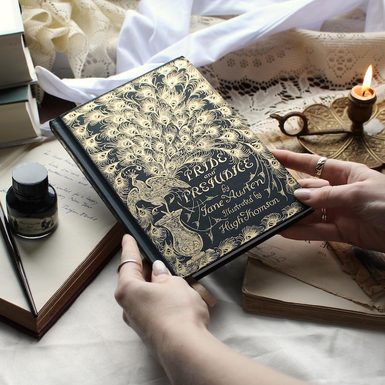 May include: A hardcover edition of "Pride and Prejudice" by Jane Austen, illustrated by Hugh Thomson. The book features a black cover with gold peacock feather and floral designs. The title is prominently displayed in elegant script.