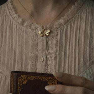 May include: A gold-colored butterfly pendant necklace. The butterfly has detailed wing patterns and is suspended from a delicate chain. The person is wearing a cream-colored blouse with lace trim and holding a book with the words "WELL READ" visible.