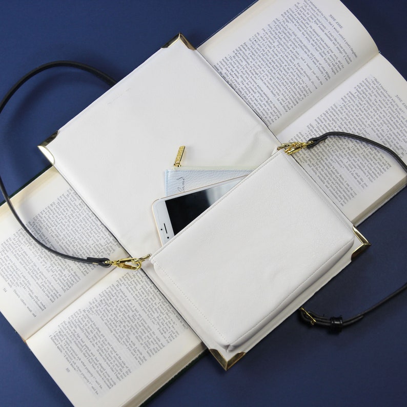 May include: A white leather crossbody bag with a gold zipper and a black strap. The bag is open and a phone and a small white pouch are inside.
