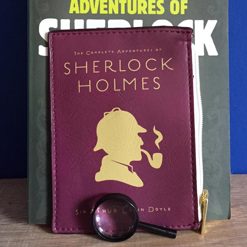 Puede incluir: Una bolsa de cremallera color burdeos con un tirador de cremallera dorado. La bolsa presenta una silueta dorada de Sherlock Holmes con una pipa. El texto "The Complete Adventures of Sherlock Holmes" est&aacute; impreso en dorado en la parte delantera de la bolsa. El texto "Sir Arthur Conan Doyle" est&aacute; impreso en dorado en la parte inferior de la bolsa.