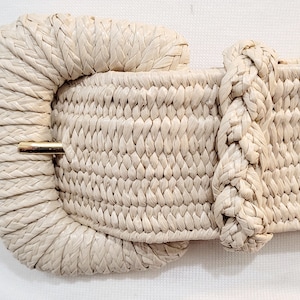 May include: A woven cream-colored belt with a large, round buckle. The buckle is also woven and has a gold metal clasp.