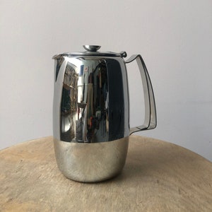 Puede incluir: Una jarra de plata con cuerpo redondeado y asa. La jarra tiene una superficie brillante y reflectante.