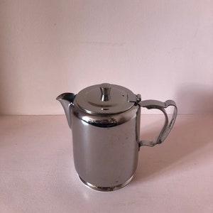 Puede incluir: Una tetera de acero inoxidable plateada con tapa y asa. La tetera está sobre una superficie blanca.