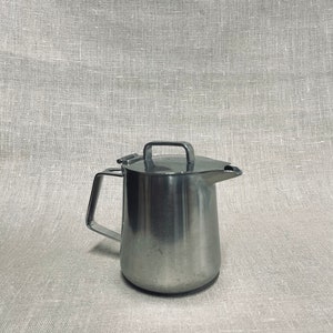 Puede incluir: Una jarra de acero inoxidable plateada con asa y pico. La jarra está sobre un fondo de lino gris claro.