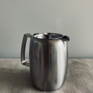 Puede incluir: Una tetera de acero inoxidable plateada con tapa y asa. La tetera está sobre un mantel de lino gris claro.