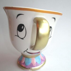 Può includere: Una tazza in ceramica bianca con un volto dipinto caratterizzato da grandi occhi e una bocca sorridente. La tazza presenta un manico color oro, un bordo dorato e una base decorata con petali alternati azzurri e rosa con finiture oro.