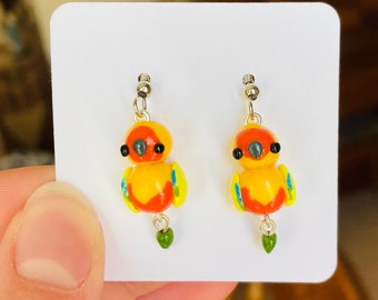 Pode incluir: Um par de brincos pendentes com pequenos pingentes de pássaros coloridos. Os pássaros são amarelos, laranja, vermelhos e verdes. Os brincos são de metal e têm um acabamento prateado.