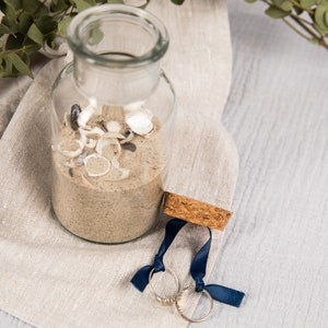 May include: A clear glass jar filled with sand and seashells, with two gold rings tied with blue ribbon on a beige linen cloth.