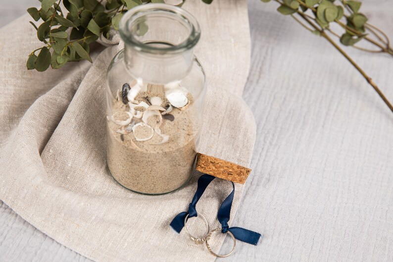 May include: A clear glass jar filled with sand and seashells, with two gold rings tied with blue ribbon on a beige linen cloth.