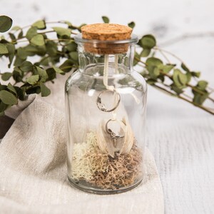 May include: A clear glass jar with a cork lid, containing two silver rings, white feathers, and dried moss. The jar is sitting on a light brown linen fabric.