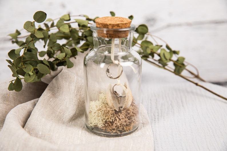 May include: A clear glass jar with a cork lid, containing two silver rings, white feathers, and dried moss. The jar is sitting on a light brown linen fabric.