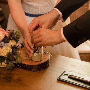 May include: A bride and groom pour sand into a glass jar during a wedding ceremony. The jar is on a wooden slice, and a bouquet of flowers is in the foreground.