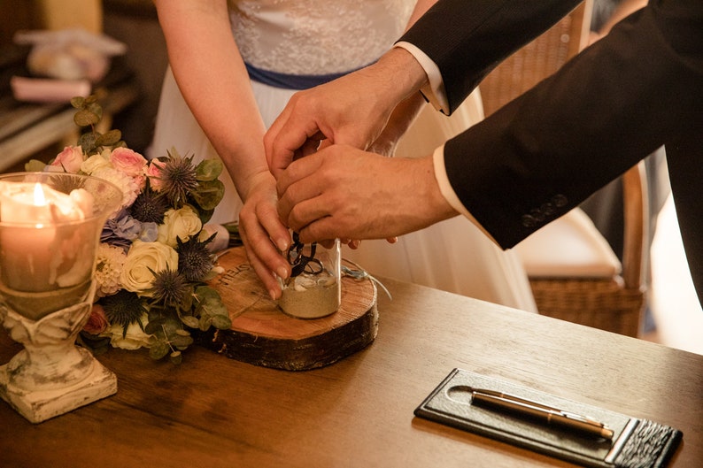 May include: A bride and groom pour sand into a glass jar during a wedding ceremony. The jar is on a wooden slice, and a bouquet of flowers is in the foreground.