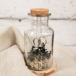 May include: Two gold wedding bands are suspended inside a clear glass jar with a cork lid. The jar is filled with dried moss and blue flowers and sits on a wooden base.