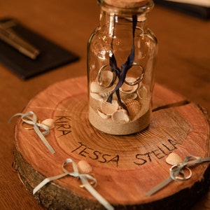May include: A wooden ring bearer box with the names "Kira", "Tessa", and "Stella" inscribed on it. The box contains a glass jar filled with sand and seashells, and three rings with blue ribbons.