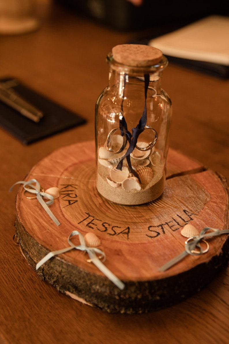 May include: A wooden ring bearer box with the names "Kira", "Tessa", and "Stella" inscribed on it. The box contains a glass jar filled with sand and seashells, and three rings with blue ribbons.