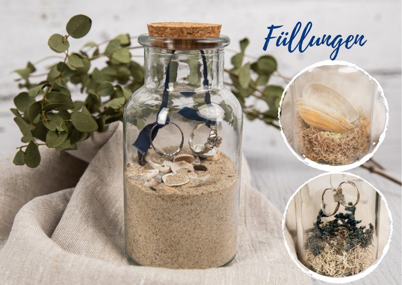 May include: A clear glass jar with a cork lid filled with sand, seashells, and two rings. The jar is sitting on a light brown fabric surface. The text "F&uuml;llungen" is visible in the upper right corner of the image.