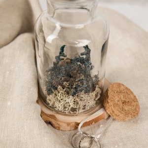 May include: Two silver wedding bands are displayed on a wooden base with a clear glass jar filled with dried moss and blue flowers. The jar has a cork stopper.