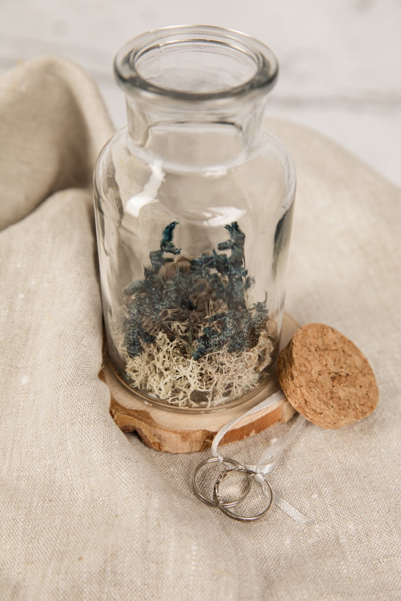 May include: Two silver wedding bands are displayed on a wooden base with a clear glass jar filled with dried moss and blue flowers. The jar has a cork stopper.