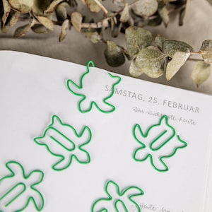 Könnte beinhalten: Fünf grüne Schildkröten-förmige Büroklammern auf einer weißen Seite mit dem Text "SAMSTAG, 25. FEBRUAR Das wichtigste heute" in schwarzer Tinte.