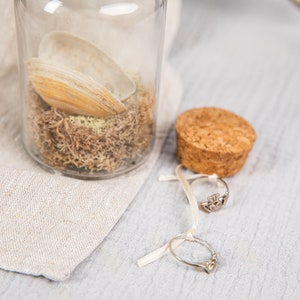 May include: A glass jar with a cork lid filled with dried moss and a seashell. Two silver rings are on a white ribbon next to the jar.