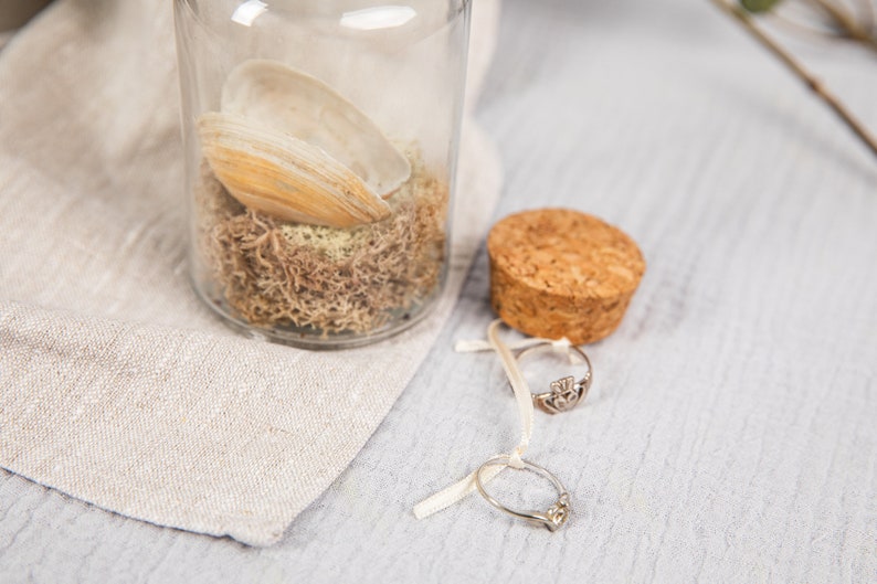May include: A glass jar with a cork lid filled with dried moss and a seashell. Two silver rings are on a white ribbon next to the jar.