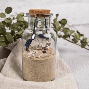 May include: A clear glass jar with a cork lid filled with sand, seashells, and two silver rings tied together with a blue ribbon.