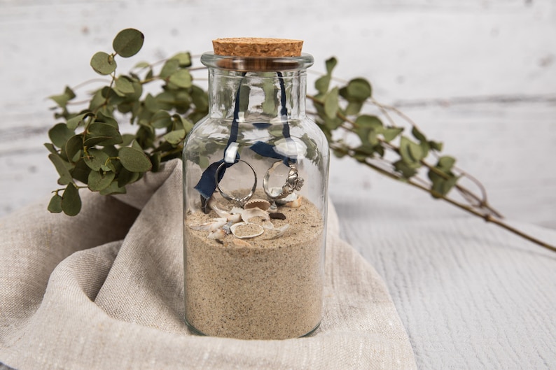 May include: A clear glass jar with a cork lid filled with sand, seashells, and two silver rings tied together with a blue ribbon.