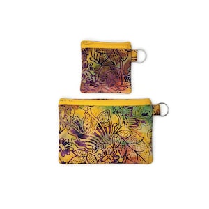 May include: Two batik fabric pouches with yellow zippers and key rings. The pouches feature a colorful floral and butterfly pattern in shades of yellow, purple, green, and orange. The larger pouch is rectangular, and the smaller one is square.