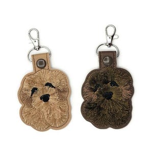 May include: Two leather keychains with embroidered dog faces. One keychain is brown with a brown dog face and the other is tan with a tan dog face.