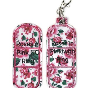 May include: Two pink and white floral patterned fabric keychains with a silver metal clasp. The keychains are shaped like a band-aid and have the text "Roses in Pink NO Ring" and "Roses in Pink with Ring" embroidered on them.