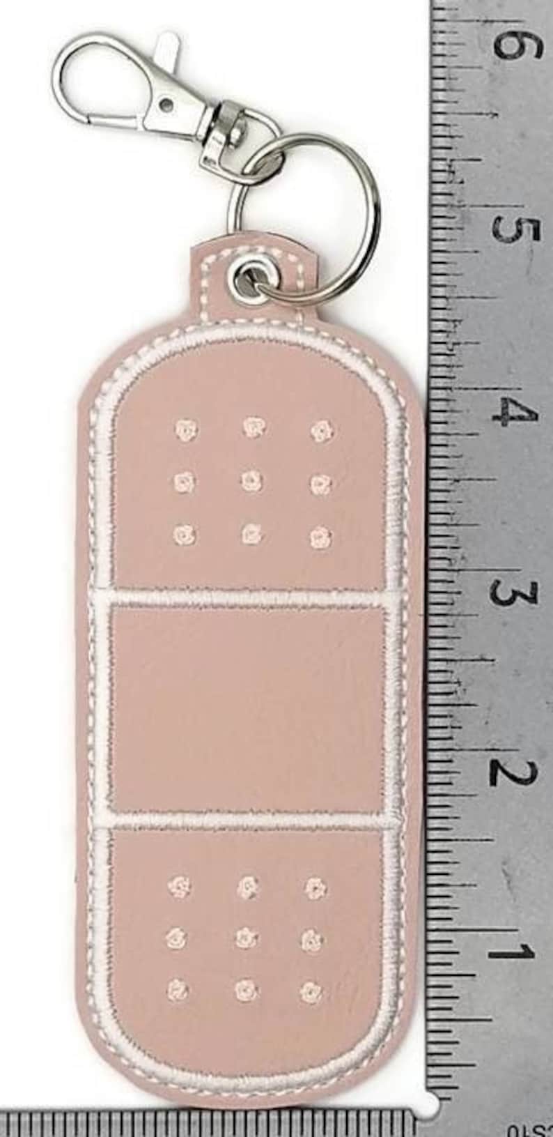 May include: A pink leather keychain shaped like a bandage with white stitching. The keychain has a silver metal ring and a silver metal clasp.