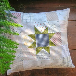 May include: Square patchwork pillow with a star design. The star is made of green and black gingham fabric. The pillow has a variety of patterned fabrics in shades of cream, red, and tan. The pillow is quilted with a ribbed texture.