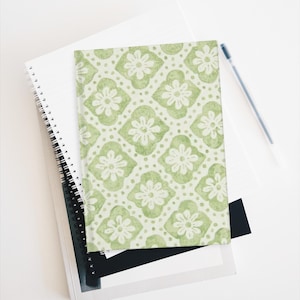 Puede incluir: Un cuaderno con un patrón floral verde y blanco, apoyado sobre una pila de otros cuadernos y un bolígrafo. La cubierta del cuaderno presenta un patrón repetido de flores estilizadas y formas geométricas.