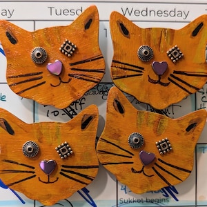 May include: Four orange wooden cat faces with black painted whiskers and eyes. Each cat face has a purple heart and two small metal embellishments on the face.