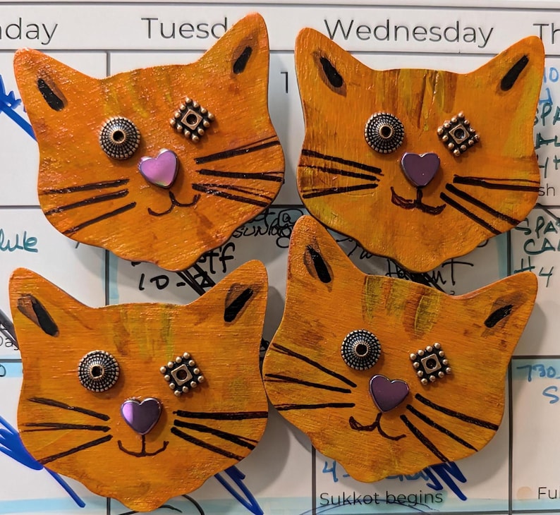 May include: Four orange wooden cat faces with black painted whiskers and eyes. Each cat face has a purple heart and two small metal embellishments on the face.